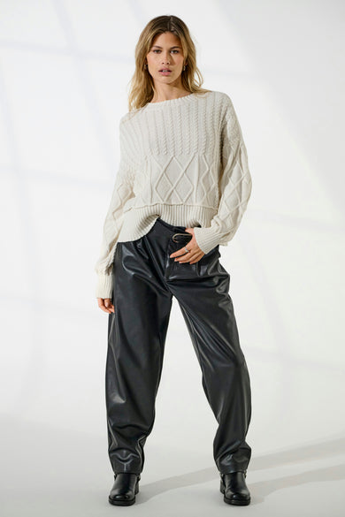 Vrouw draagt witte trui, zwarte leren broek en zwarte laarzen, staand.
