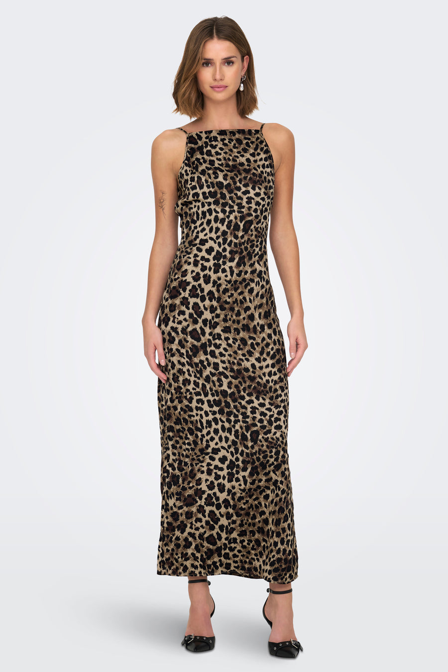 Vrouw in luipaardjurk met zwarte hakken, staand tegen witte achtergrond.