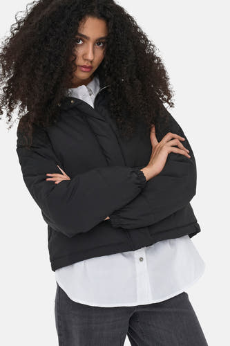 Manteau court noir de JDY, avec des manches longues et porté sur une chemise blanche et un jean gris.