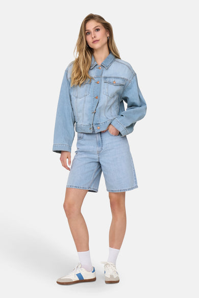 Lichtblauwe denim short van ONLY, gedragen met een denim jas en witte sportschoenen.
