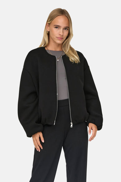 Zwarte korte jas van ONLY, met een zilverkleurige rits en gedragen met een grijze top en zwarte broek.
