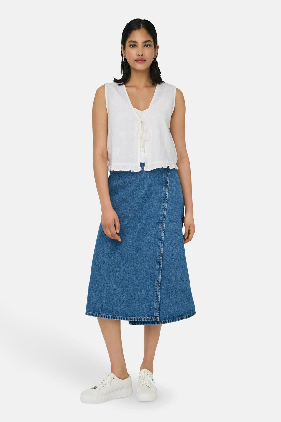 Witte mouwloze blouse van ONLY, met bloemenborduurwerk en koordsluiting vooraan, gecombineerd met een denim rok en witte sneakers.
