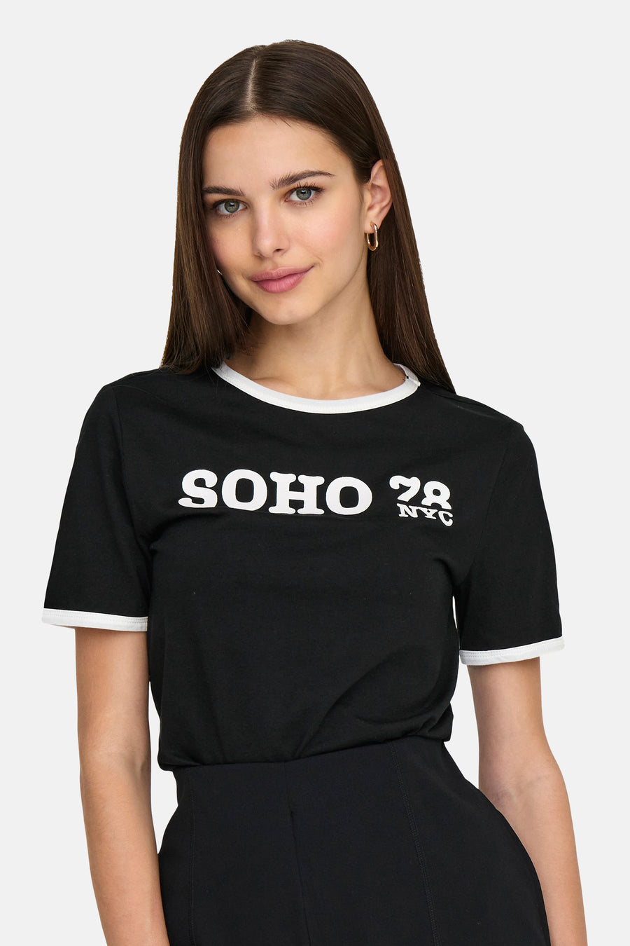 T-shirt noir à manches courtes de ONLY, avec un bord blanc et 'SOHO 78 NYC' imprimé, porté avec une jupe noire et des boucles d'oreilles dorées.