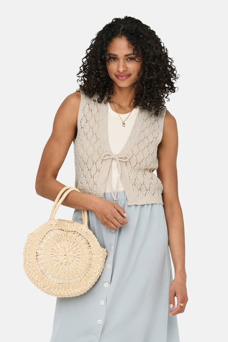 Ecru cardigan van ONLY, met opengewerkt breipatroon, striksluiting en gecombineerd met een blauwe rok en een rieten tas.
