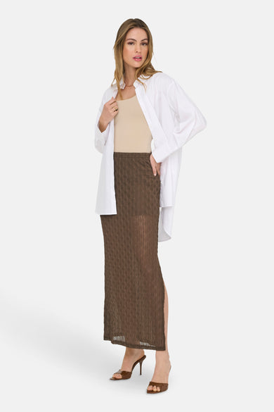 Lange bruine rok van ONLY, met een subtiele textuur en een hoge split, gecombineerd met een beige top en een wit overhemd.
