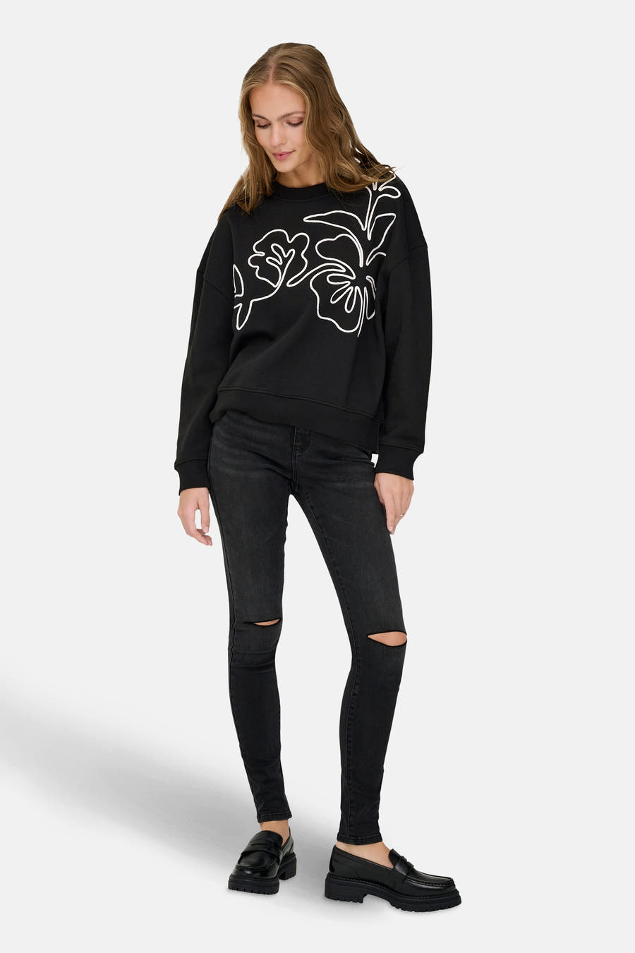 Zwarte sweater met ronde hals van JDY, met een witte bloemenprint, gedragen met zwarte jeans met gaten en zwarte loafers.
