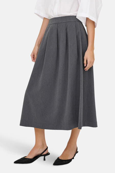 Jupe grise de JDY, avec des plis et une longueur jusqu'aux genoux, portée avec une blouse blanche et des bottines noires.