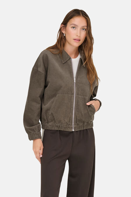 Bruine korte jas van ONLY, met zilveren ritssluiting en gedragen met een wit shirt en een bruine broek.
