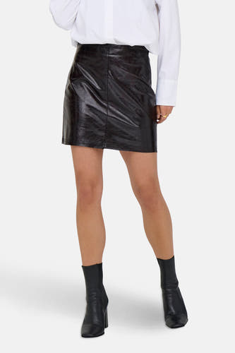 Jupe brune courte de JDY, avec une texture brillante et en cuir et associée à des bottes noires et un chemisier blanc.
