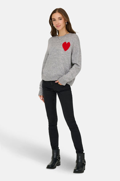 Grijze ronde hals trui van JDY, met rood hart op de borst, gecombineerd met zwarte jeans en laarzen.
