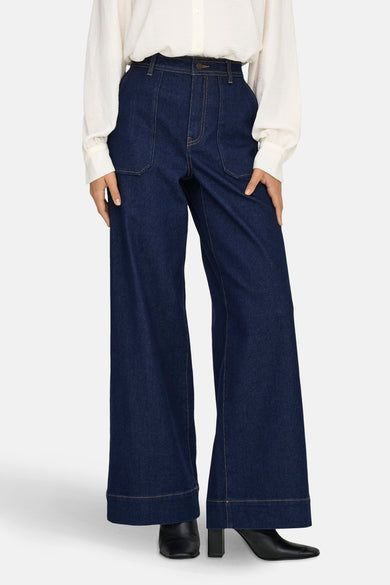 Jean large en denim bleu foncé de JDY, avec des jambes larges, sur un modèle avec une blouse blanche.
