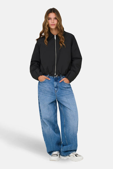 Veste courte noire de ONLY, à manches longues, avec une fermeture éclair argentée, portée avec un jean ample et des baskets.