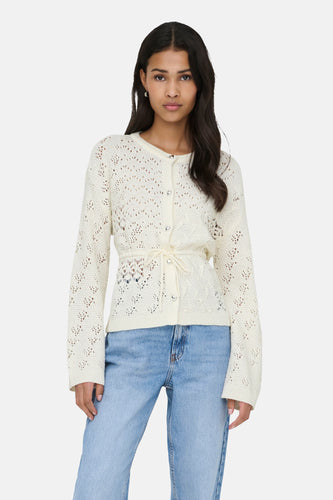 Vrouw in een gebreid, ecrukleurig vest met een tailleceintuur en blauwe jeans tegen een witte achtergrond.