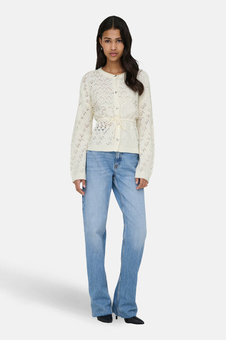 Een vrouw in een witte cardigan en blauwe jeans, staand.