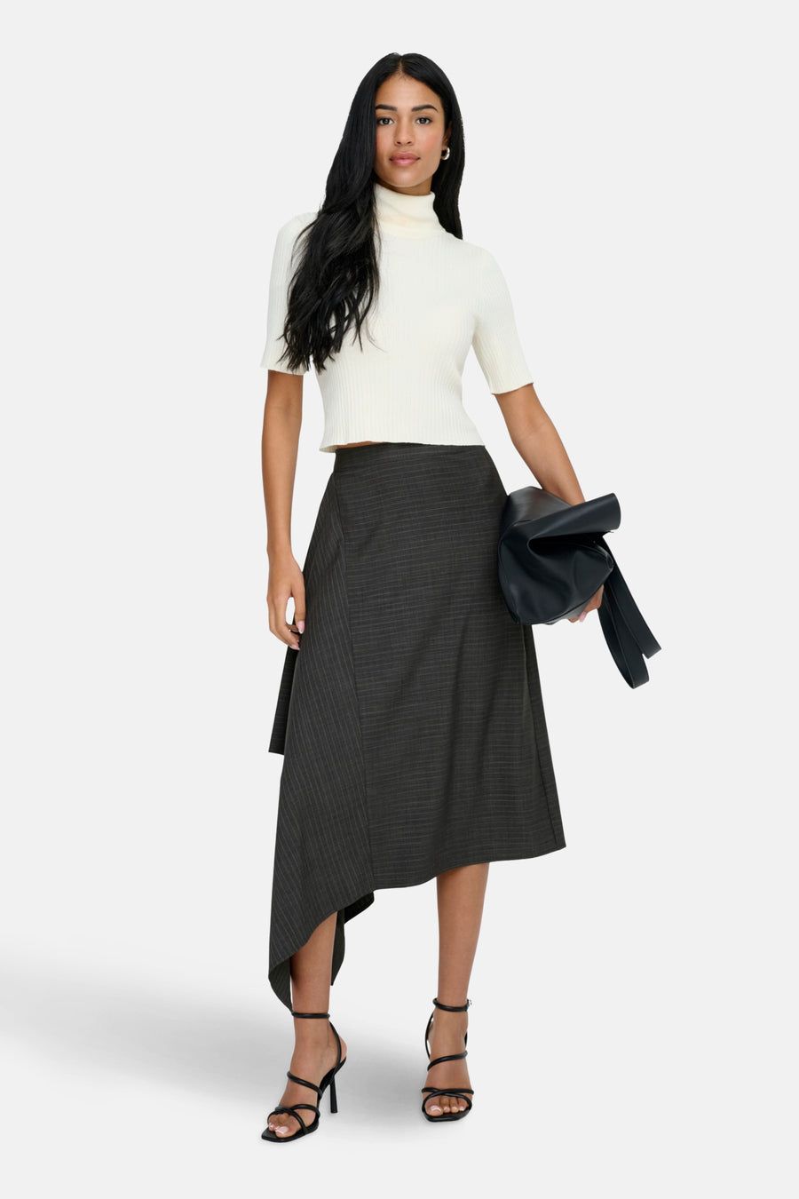 Vrouw in witte coltrui, grijze rok en zwarte sandalen met een zwarte tas in haar hand.