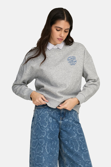 Grijze sweater van ONLY met lange mouwen, afgewerkt met een blauwe print en gedragen met een wit overhemd en blauwe broek.
