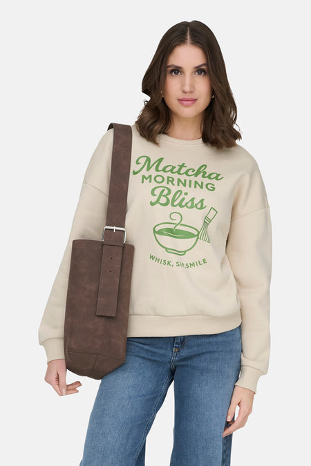 Ecrukleurige sweater met ronde hals van ONLY®, met groene "Matcha Morning Bliss" print, gedragen met jeans en bruine tas.