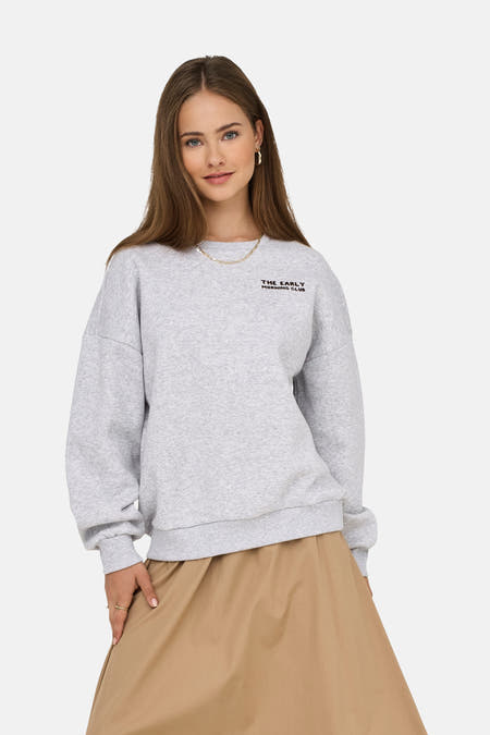 Pull-over gris d'ONLY, avec une impression textuelle sur la poitrine, porté avec une chaîne dorée et une jupe beige.