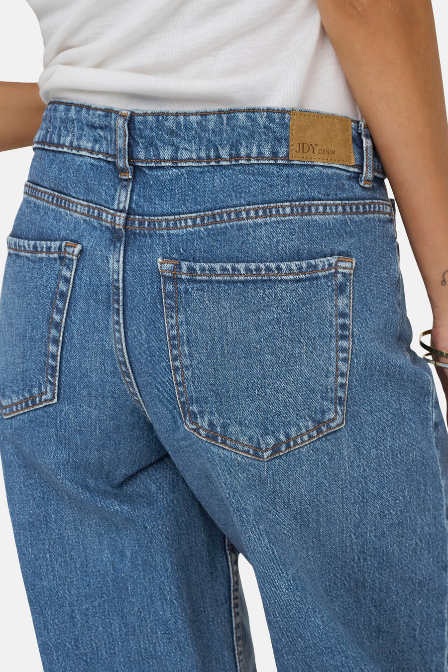 Jean large en denim bleu moyen de JDY, vue de dos, avec patch en cuir marron, coutures orange et poches arrière, porté avec un t-shirt blanc.