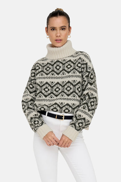 Vrouw in een beige en zwart gebreide trui met geometrische patronen, witte broek en zwarte riem met gouden gesp.