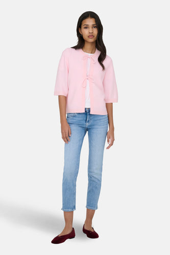 Een staande vrouw in een roze vest, een wit shirt, een blauwe jeans en rode ballerina's.