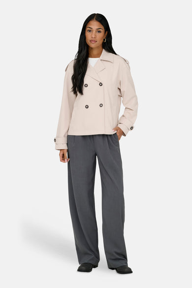 Beige jas van ONLY, met dubbele knoopsluiting, gedragen over een wit shirt en grijze pantalon.
