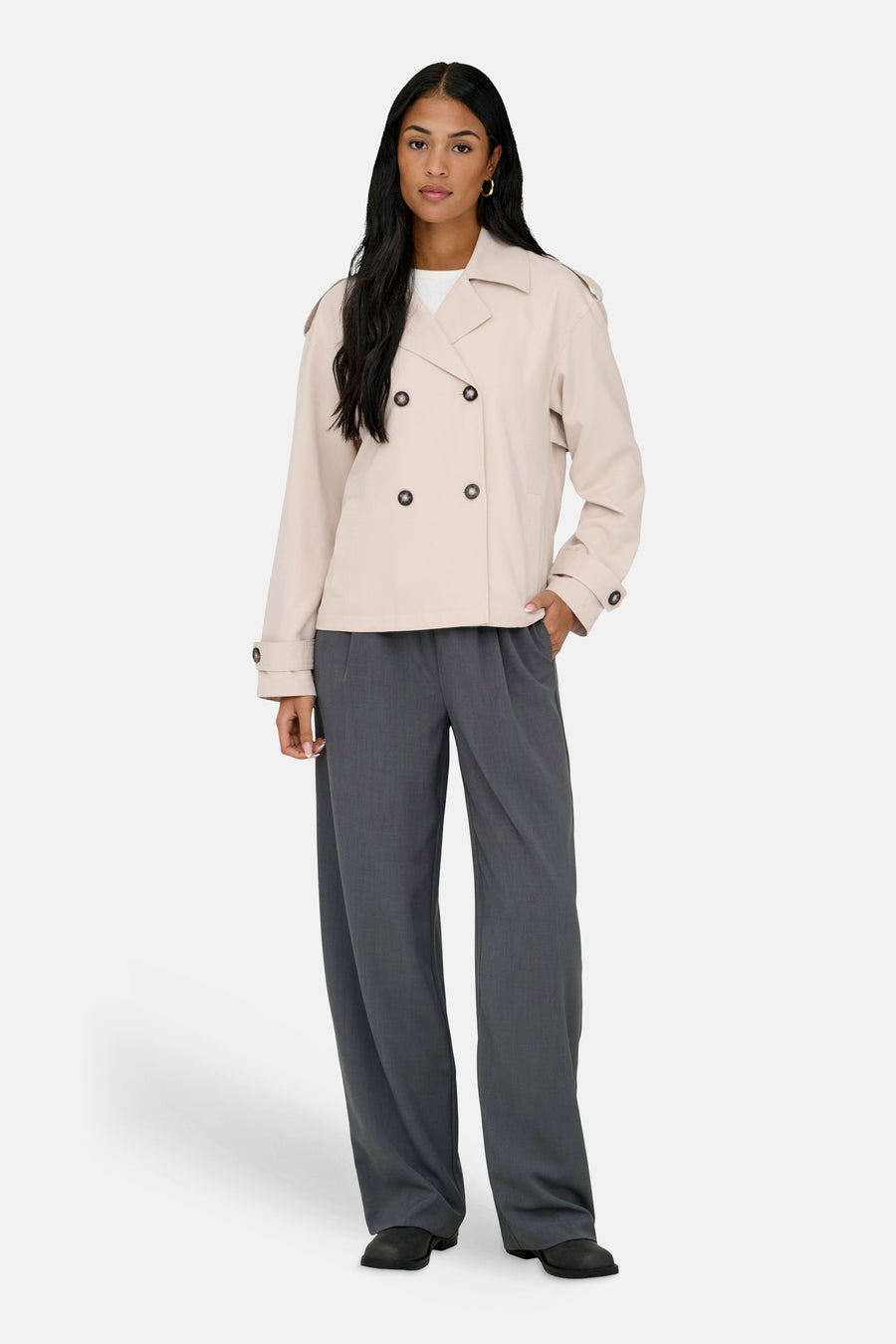 Beige jas van ONLY, met dubbele knoopsluiting, gedragen over een wit shirt en grijze pantalon.
