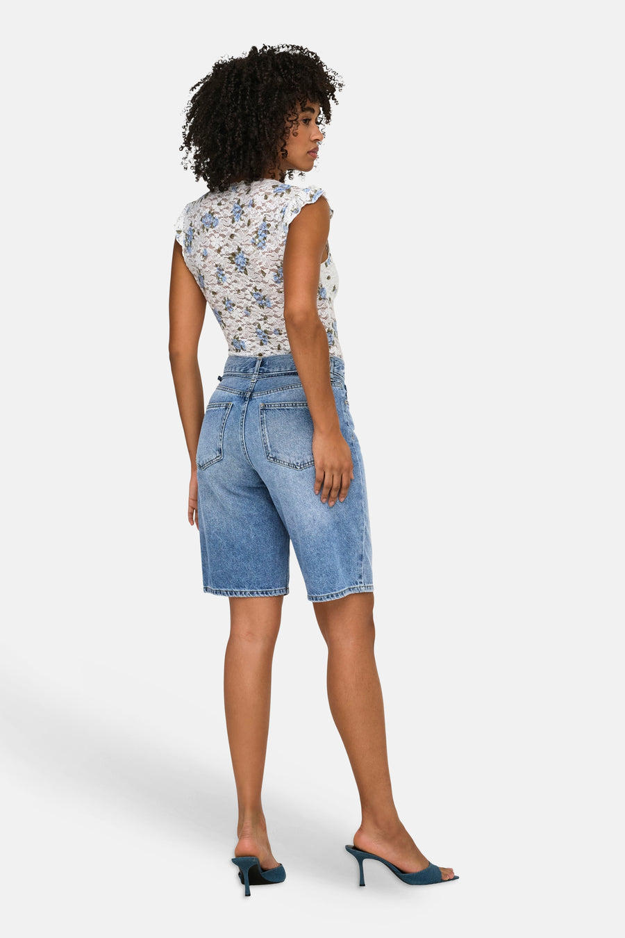 Short en jean bleu moyen de ONLY®, partiellement vue de dos sur un mannequin, combinée avec un haut fleuri en dentelle blanche et des mules bleues.