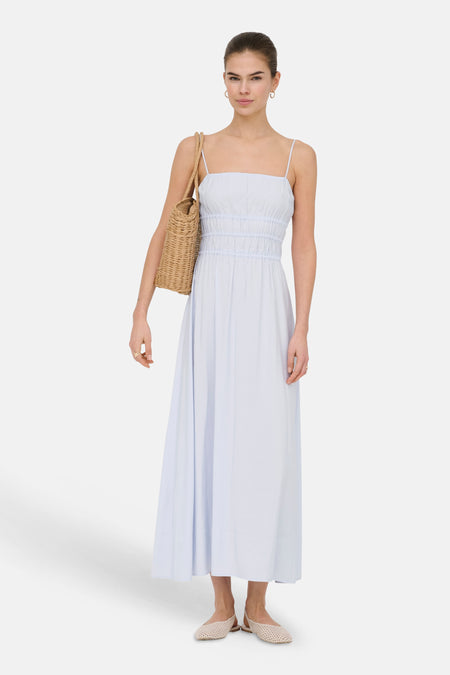 Lichtblauw maxi-kleedje van ONLY met spaghettibandjes en gesmokte taille, gestyled met een rieten tas en beige platte schoenen.