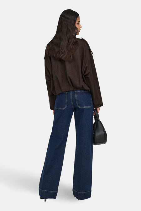 Een vrouw in een bruine blouse en wijde, donkerblauwe jeans, van achteren gezien, met een zwarte handtas.