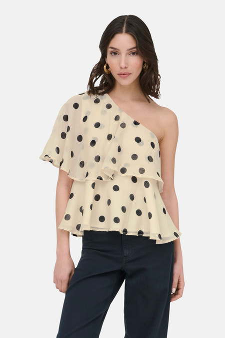 Ecru blouse van JDY met zwarte stippen, een one-shoulder ontwerp met ruches, gedragen met een donkere broek.