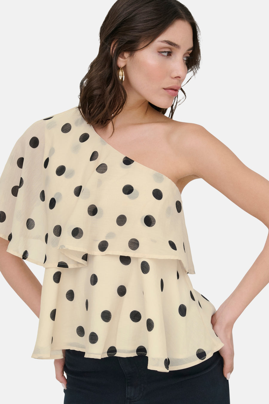 Blouse écru à manches courtes de JDY avec des pois noirs, un design asymétrique et des volants superposés, portée par un mannequin avec un pantalon noir.