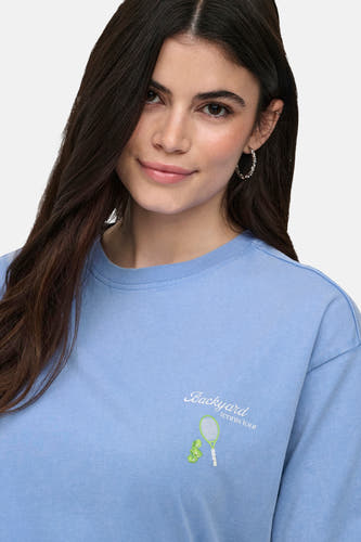 T-shirt bleu ONLY® avec inscription « Backyard tennis tour » et imprimé raquette et balles de tennis sur la poitrine, porté par un mannequin souriant.