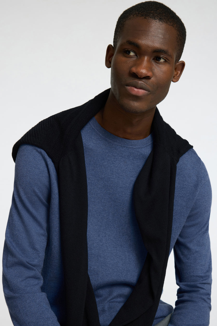 Pull bleu à col rond de SELECTED, avec un pull noir négligemment jeté sur les épaules.