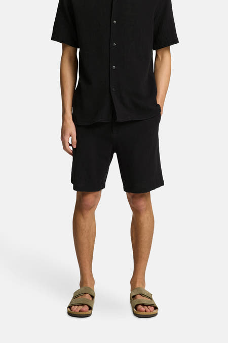 Zwarte short van SELECTED, gedragen met een zwart overhemd en bruine sandalen.
