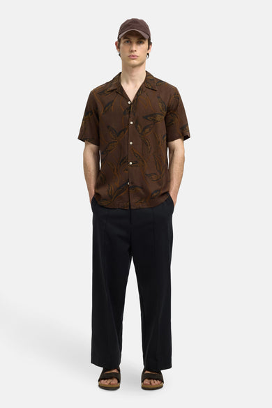 Chemise brune à manches courtes de SELECTED, imprimée de feuilles, portée avec un pantalon noir et des sandales.