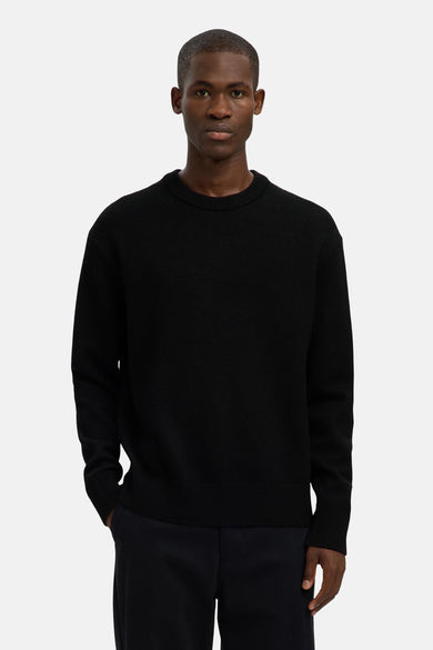 Pull-over noir avec col rond de SELECTED, porté par un homme, avec une main dans la poche.
