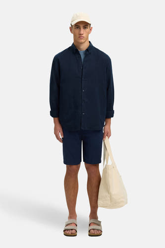 Blauw overhemd met lange mouwen van SELECTED, met opgerolde mouwen, gedragen met blauwe shorts en sandalen, met een crème katoenen tas.
