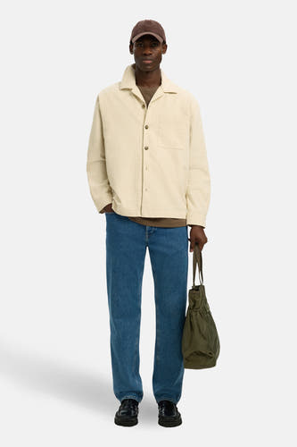 Chemise écrue de SELECTED, à manches longues et avec une poche sur la poitrine, portée avec un jean et une casquette marron.