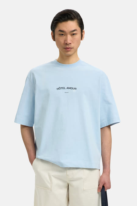 Blauw T-shirt van SELECTED, met korte mouwen en de tekst "Hôtel Amour", gedragen met een crèmekleurige broek.
