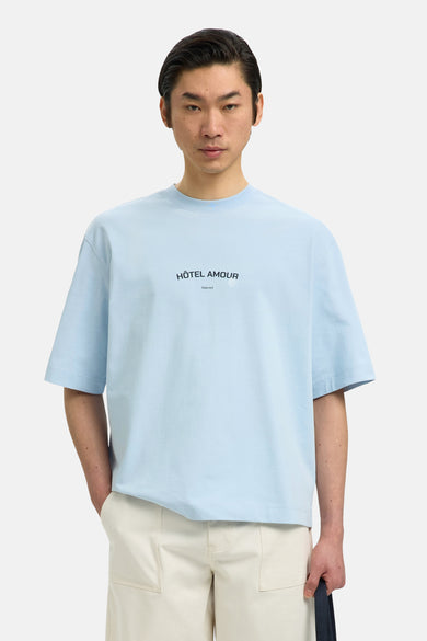 T-shirt bleu de SELECTED, à manches courtes et avec le texte "Hôtel Amour", porté avec un pantalon de couleur crème.