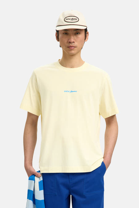 Ecru T-shirt van SELECTED met een klein blauw logo, gedragen met blauwe broeken en een witte pet.
