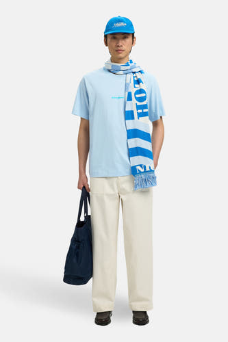T-shirt bleu (manches courtes) de SELECTED, avec une teinte bleu clair et un imprimé texte subtil, porté avec une écharpe rayée et une casquette bleue.