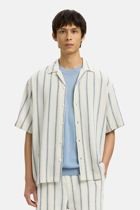 Chemise écru à manches courtes de SELECTED, rayée et portée sur une chemise bleue, avec short et sac assortis.