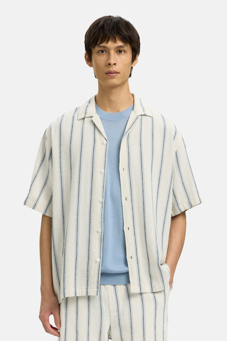 Chemise écru à manches courtes de SELECTED, rayée et portée sur une chemise bleue, avec short et sac assortis.