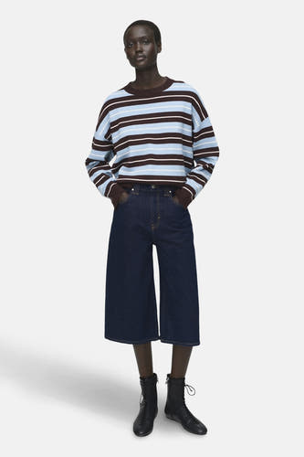 Sweat-shirt multicolore à col rond de MANGO, avec des rayures horizontales en brun, bleu et blanc, associé à un pantalon de denim large et des baskets noires.