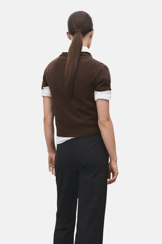 Bruine trui met V-hals van MANGO, met korte mouwen en een witte blouse eronder, gecombineerd met een donkere pantalon, gezien van achteren.
