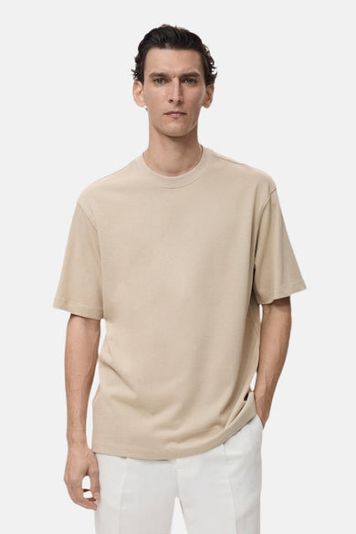 T-shirt beige de MANGO, porté avec un short blanc et posant décontracté contre un fond uni.