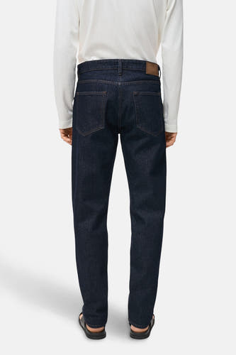 Donkerblauwe denim jeans van MANGO, gezien van achteren, gecombineerd met een wit shirt met lange mouwen en zwarte sandalen.
