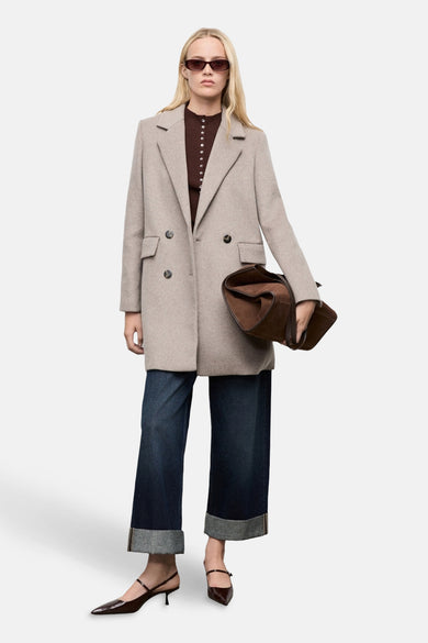 Manteau long marron de MANGO, avec des boutons doubles, porté avec un gilet marron, des jeans larges et des baskets.